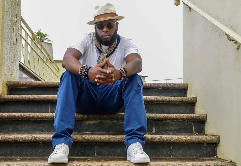 Rapper Talib Kweli sits on outdoor stairs wearing a fedora, sunglasses, white t-shirt, blue pants, and white sneakers with hands clasped.